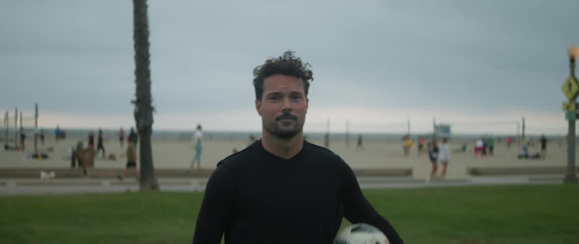 A man is standing in a field with his foot on a soccer ball as he lifts it with his foot and grabs the ball with his hands