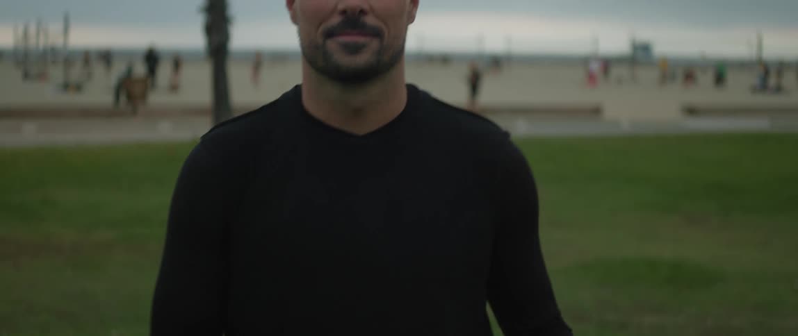 A man is standing in a field bouncing a soccer ball into the air with his feet and he catches the ball in his hand and smiles