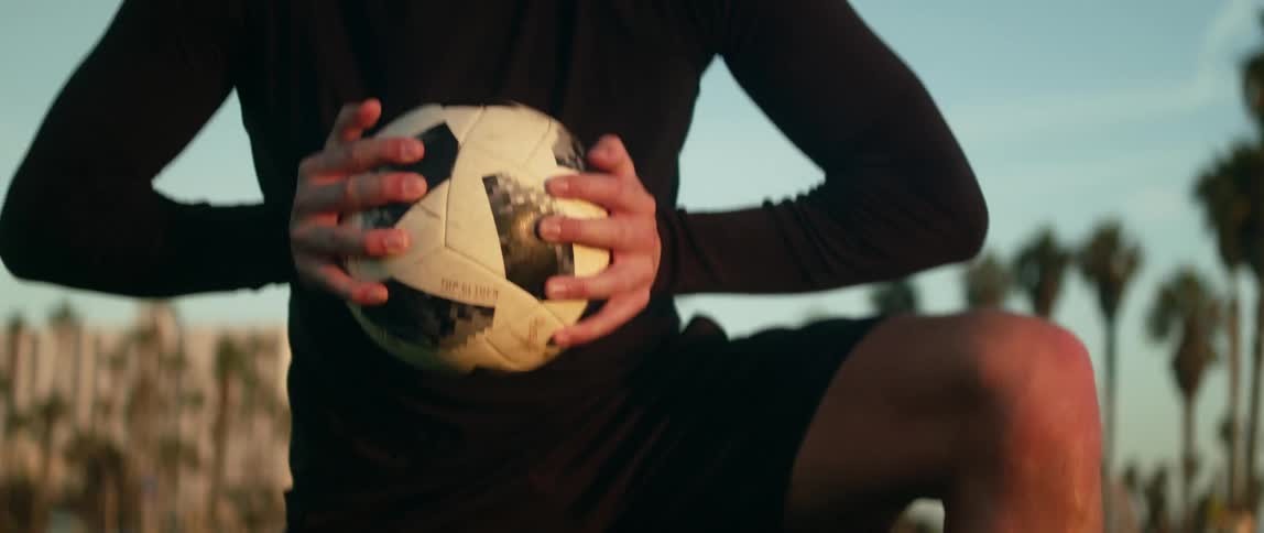 A man kneeling on a field picks up a soccer ball and squeezes it