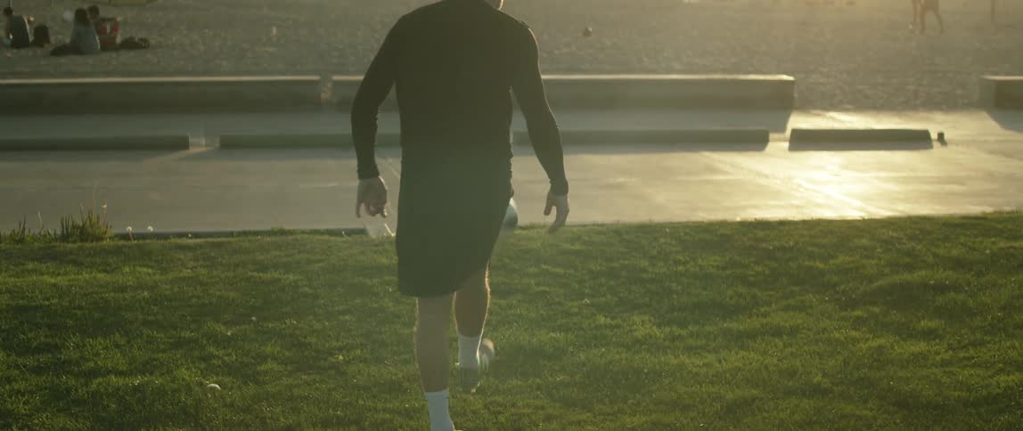 A man kicks a soccer ball in a public park by a beach where people are sitting in the sand