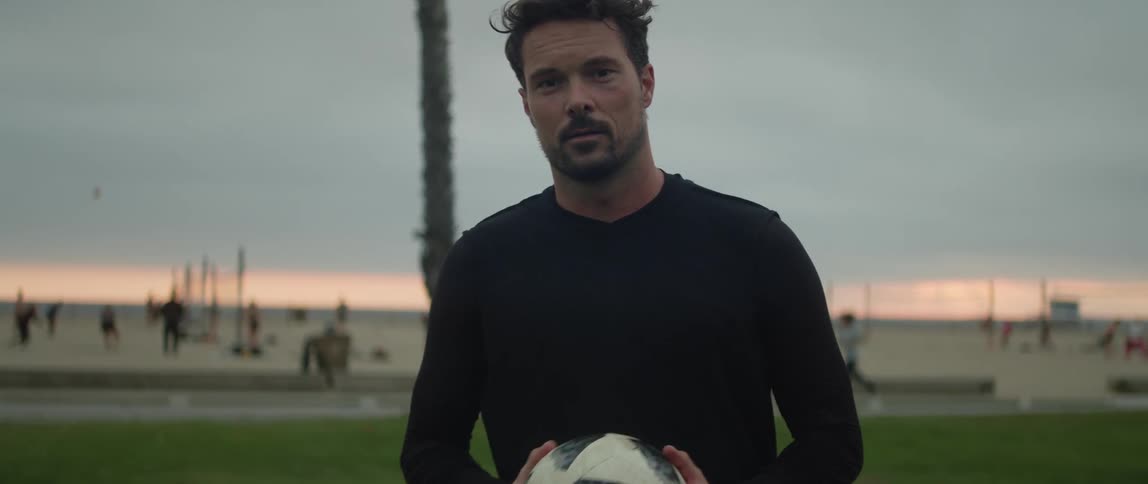 A man is standing in the grass at a beach holding a soccer ball