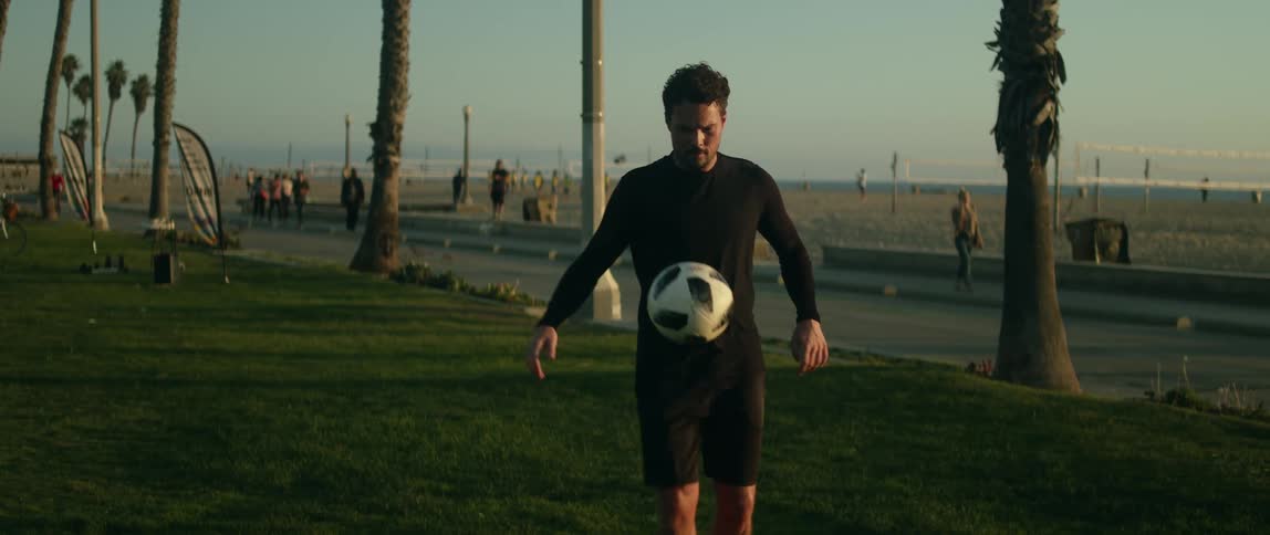 Man with black sweatshirt and shorts juggles a soccer ball on green lawn near the beach at sunset