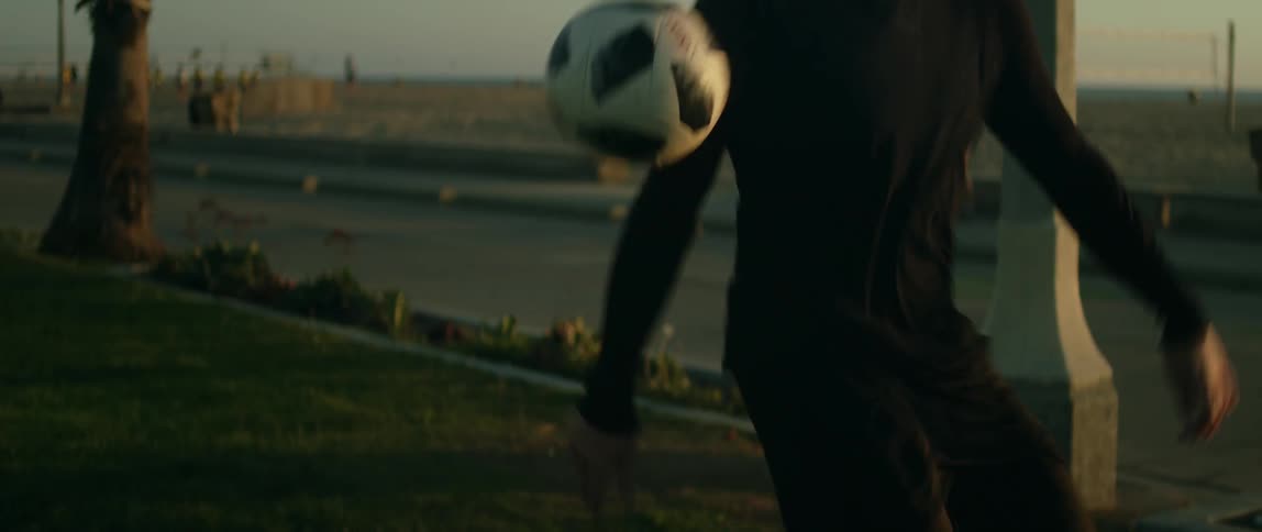 A man is standing in a park and he kicks a soccer ball so it lands in front of him as people walk in the distance