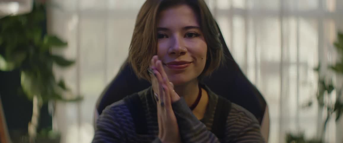 A teenage girl sits at home and smiles and puts her hands together and rests her face on them