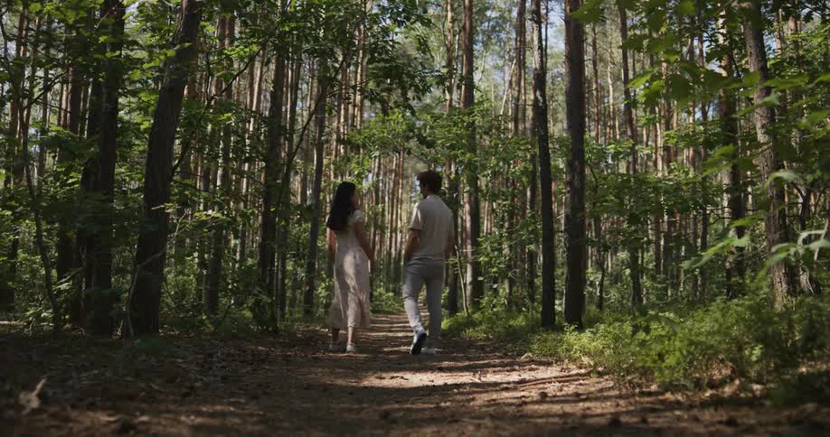 A man and a woman are walking on a path through the woods as the leaves on trees move in the breeze