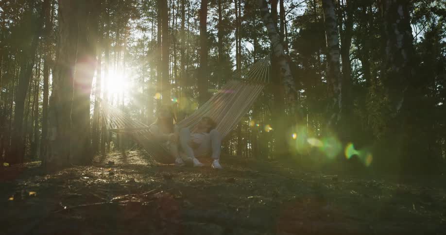 The sun shines brightly through the trees in the woods as a man and a woman relax together on a hammock