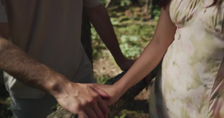 A teenage couple is in the woods holding hands atop a downed tree trunk and rocking it back and forth