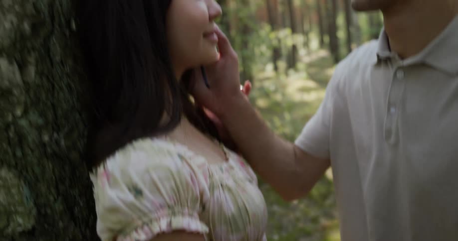 Man holds womans hand her waist and strokes her face as they stand near tree in woods on a sunny day
