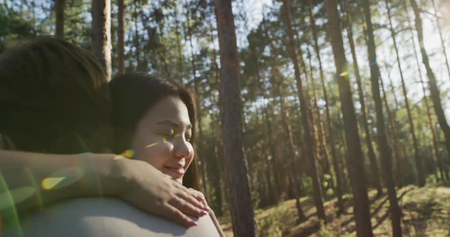 A teenage girl has her arms around her boyfriend as the sun shines through the trees in the woods