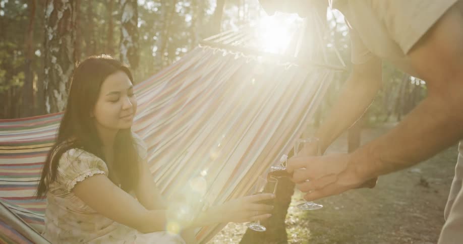 A young woman sits on a hammock in the woods as her boyfriend pours them both a glass of wine
