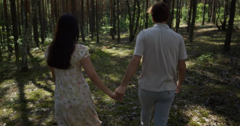 A man and a woman are running and playing around the trees in the woods and he catches her and they walk away holding hands