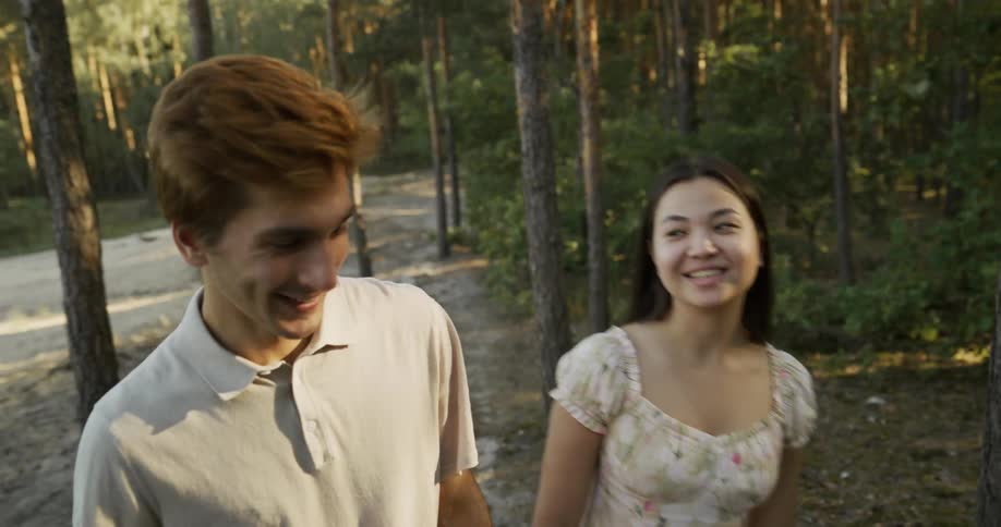 A teenage couple walk through a wooded area holding hands laughing and talking