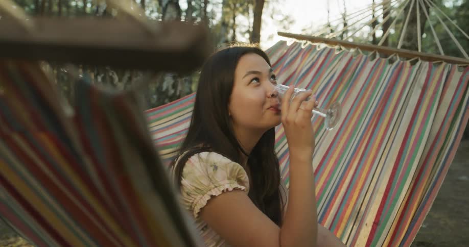 A woman is sitting on a hammock in the woods drinking a glass of wine with a smile on her face