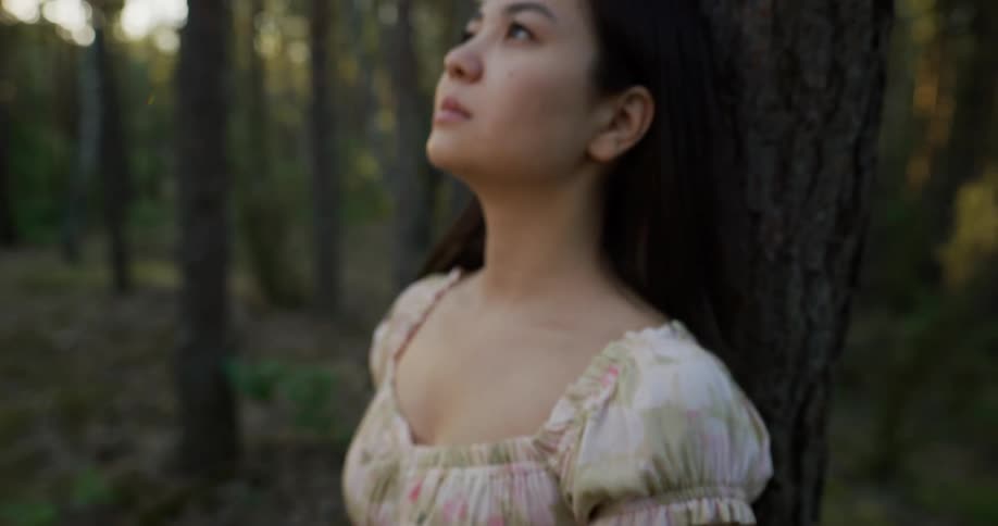 A woman wearing a white flowered dress is standing in the woods leaning against a tree