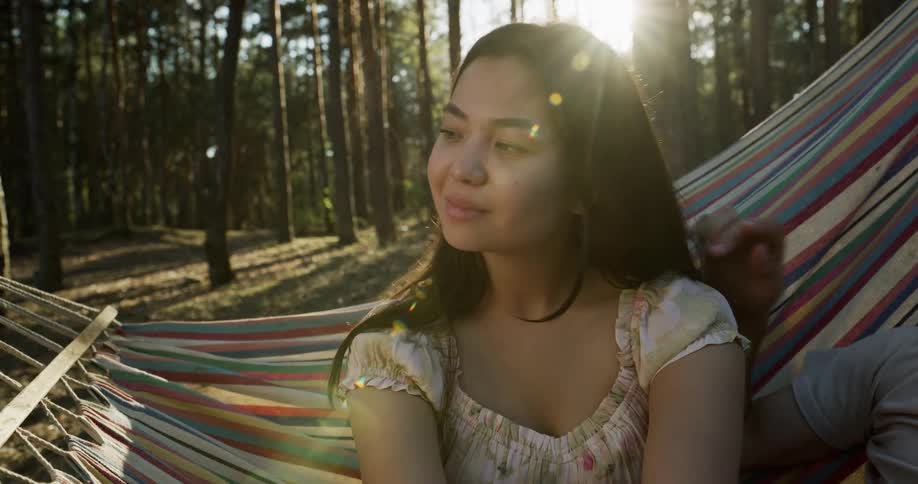 A teenage girl is sitting on a hammock as someone moves her hair with their hand and the sun shines through the trees in the woods