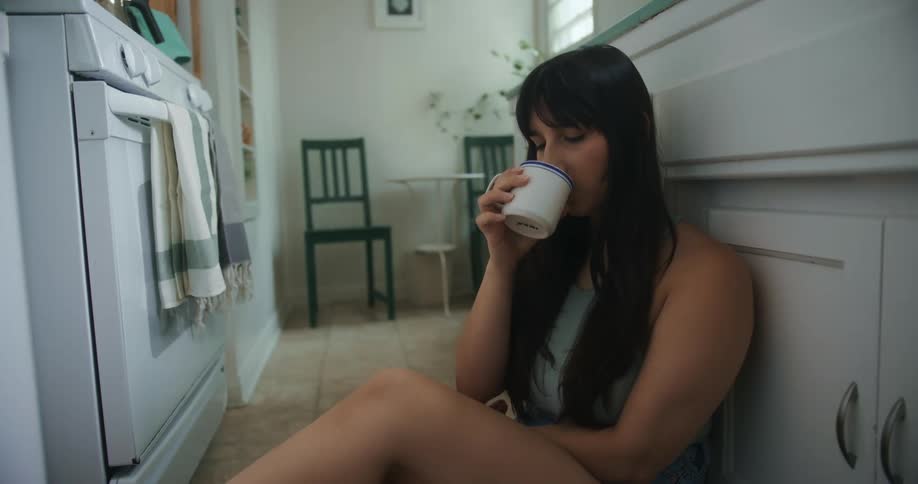 Woman sits on floor in kitchen drinking from mug
