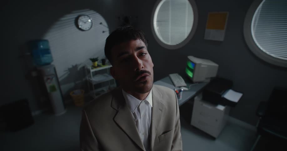 A man standing in an office chewing gum with and old computer and printer on a desk behind him