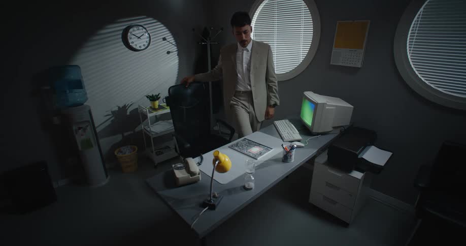 A man staring out a round window checks his watch then sits back down at his desk