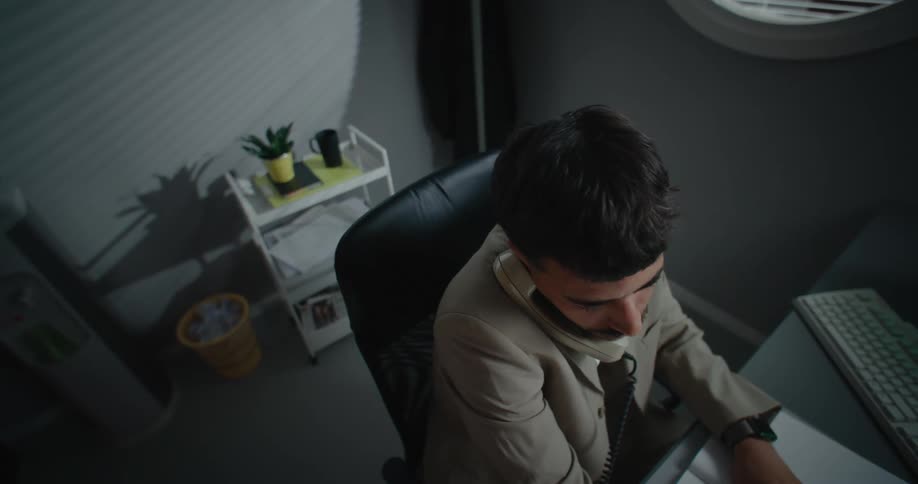 A man is sitting in a chair in front of a desk with a computer and he answers the phone and opens a book as he talks and he hands up the phone and tears a page out of the book and balls it up and throws it over his shoulder