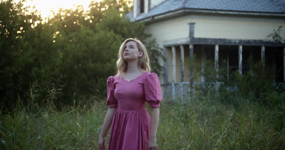 A woman walks through tall grass toward an abandoned home