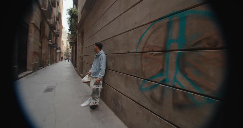 A man is standing in an alley leaning against a building and holding his skateboard