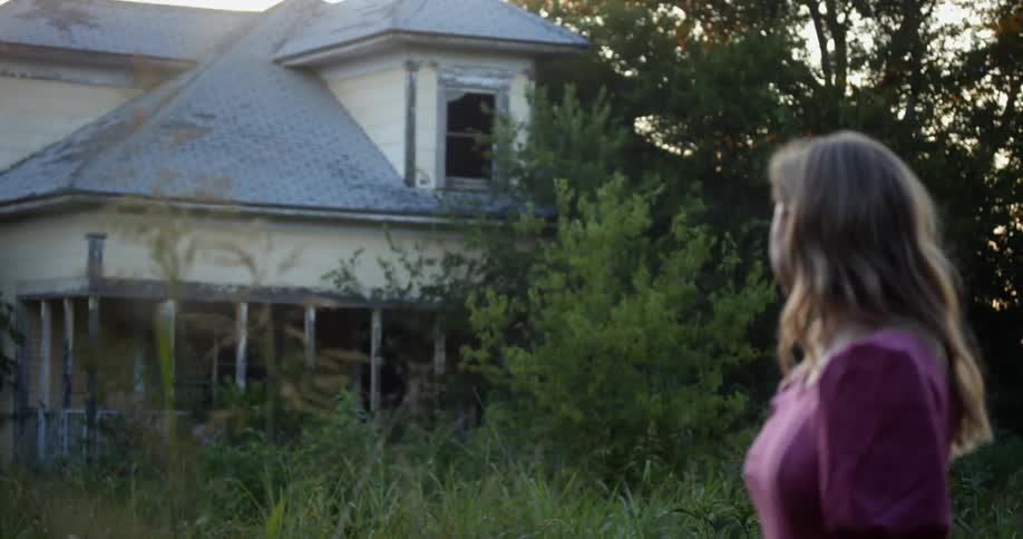 A blond woman in a pink dress is standing outside of an abandoned house with an overgrown unkempt yard