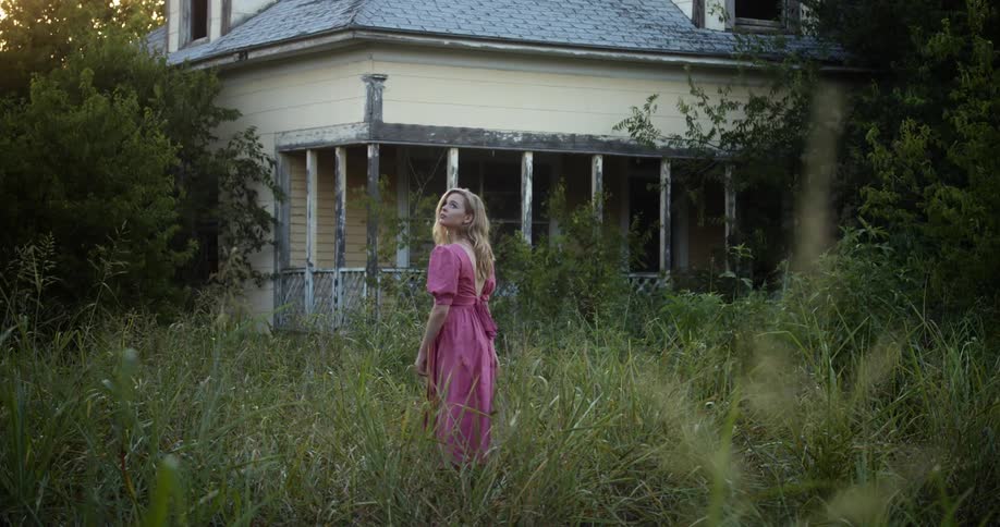 A young woman walks through tall grass towards a house in a rural area