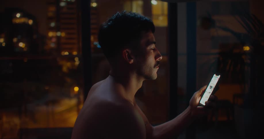 A man is sitting in his room holding his cell phone and he falls back onto his bed as he looks at it