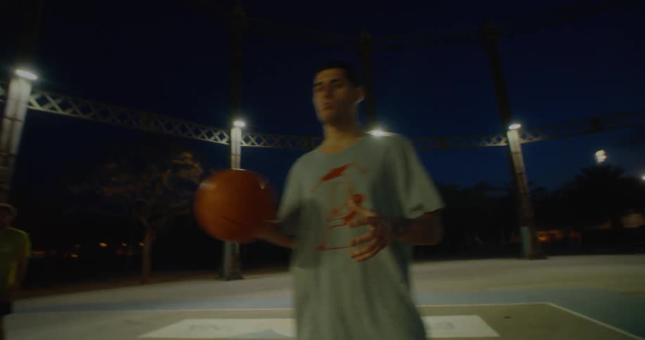 A man catches a passed basketball on an outdoor court at night