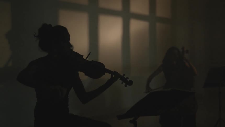 A woman is playing a violin and another woman is playing a big bass guitar in a dark room