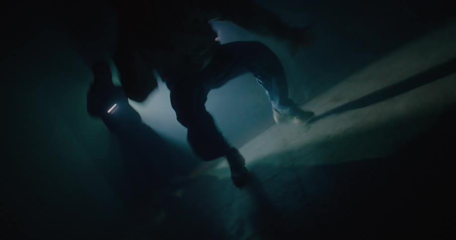 A man is break dancing in a dark room as someone behind him holds a flashlight and someone else walks around