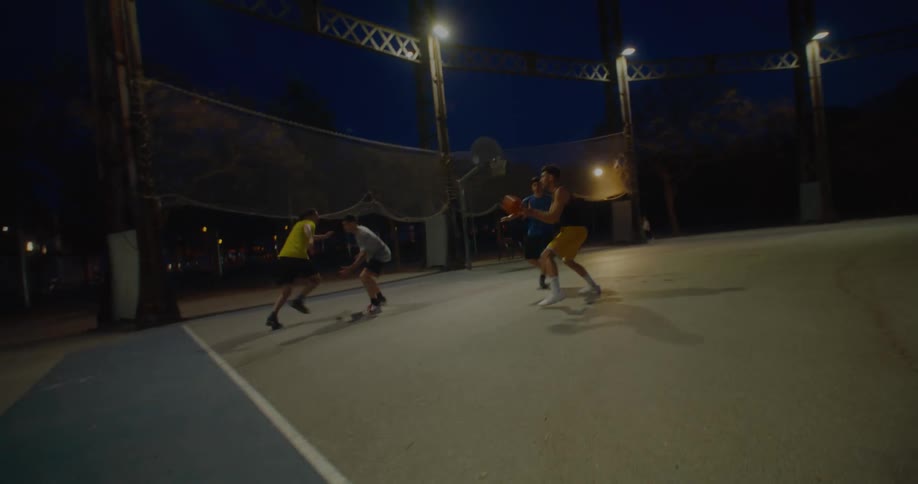 A group of four friends playing two on two basketball on an outdoor court at night