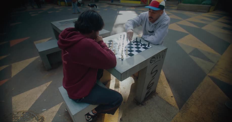 Two men are sitting down at a table in a park playing chess