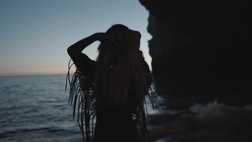 Woman runs her fingers through her hair by the ocean shore at dusk