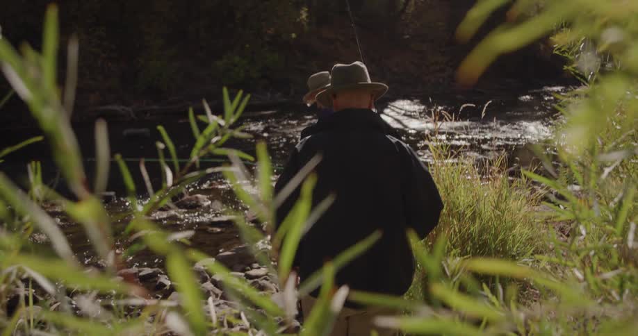 Two men wearing hats are fishing in a stream in the woods