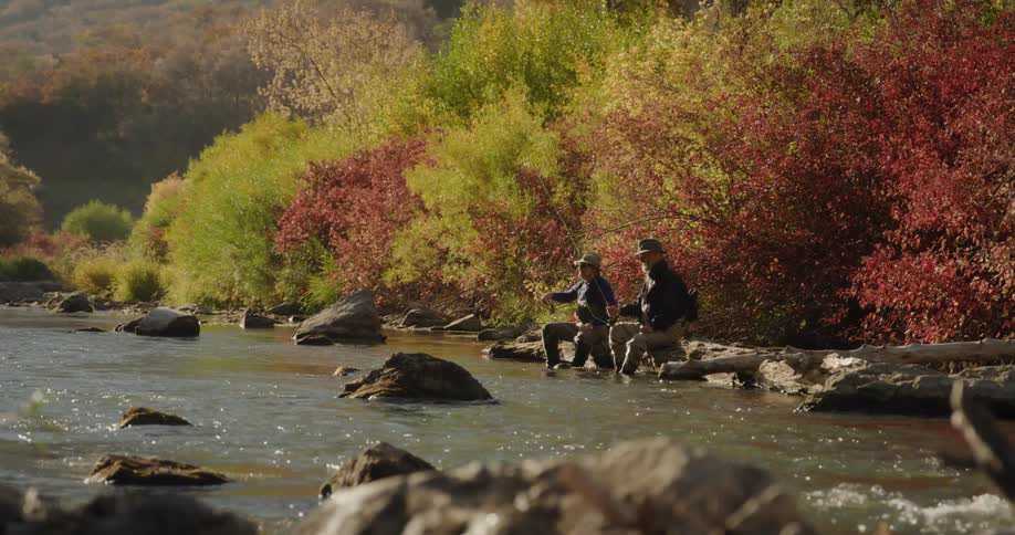 Two friends wearing hats sunglasses jacket and pants sits on rock side by side fish in a river near the forest with brown trees
