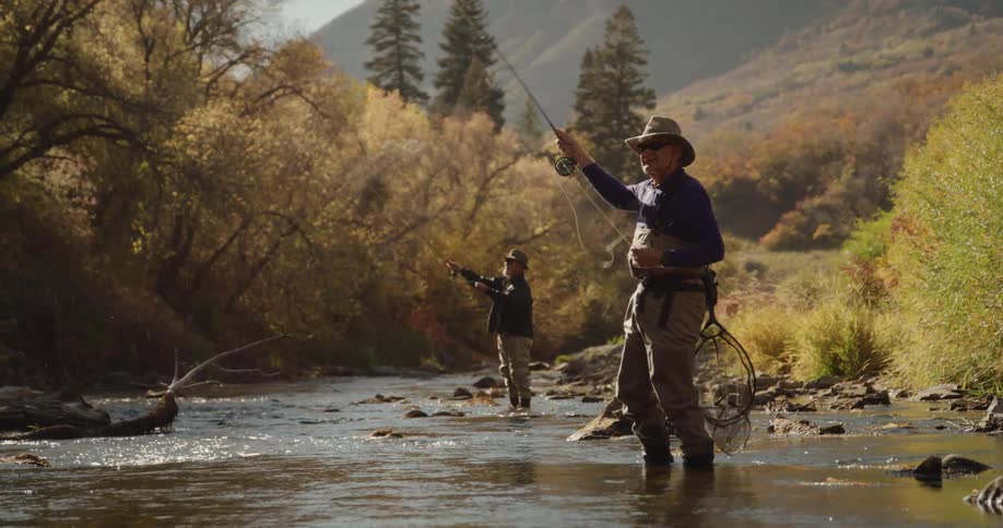 Two men are standing along a riverbank near the woods casting their fishing lines into the water