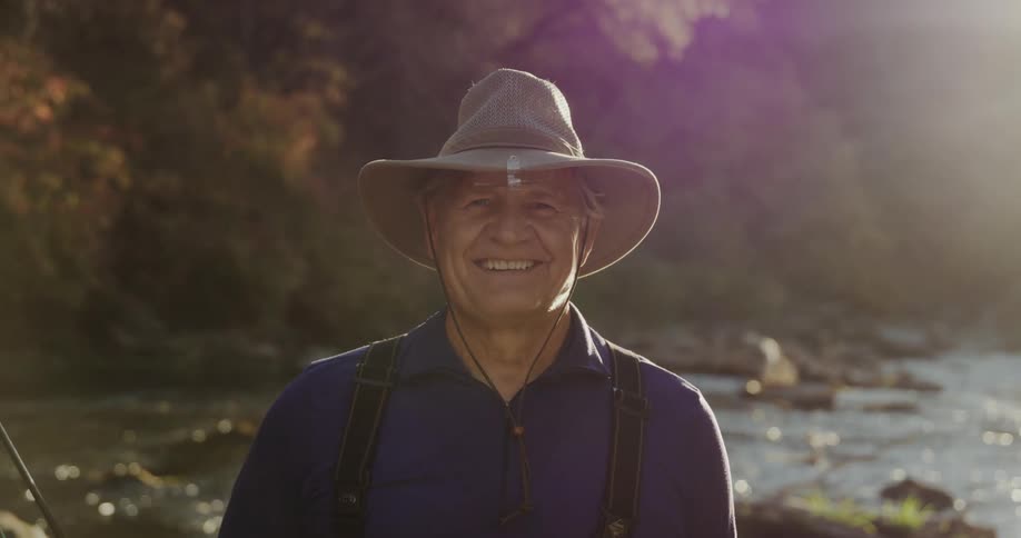 Man stands outdoors near stream on bright day holding fishing rod and smiling