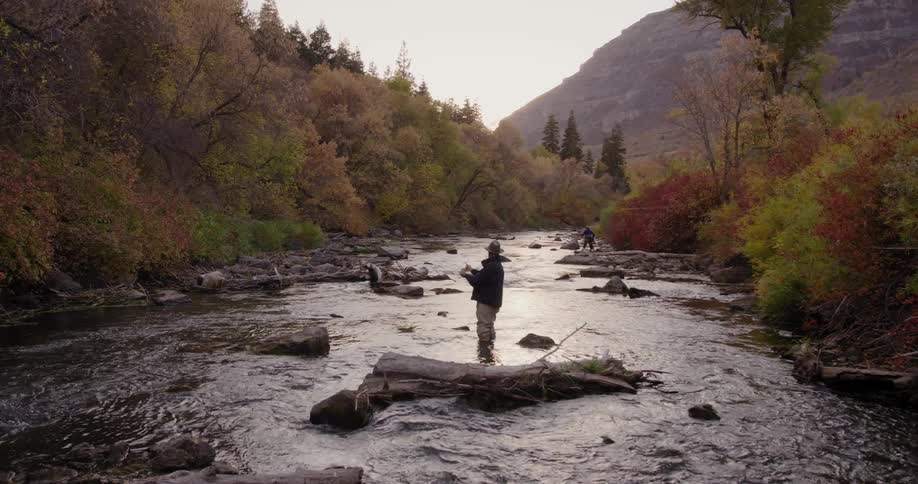 Two men wearing hats sunglasses and black coat and brown pants cast fishing rods in the forest river near brown trees with mountain in the background