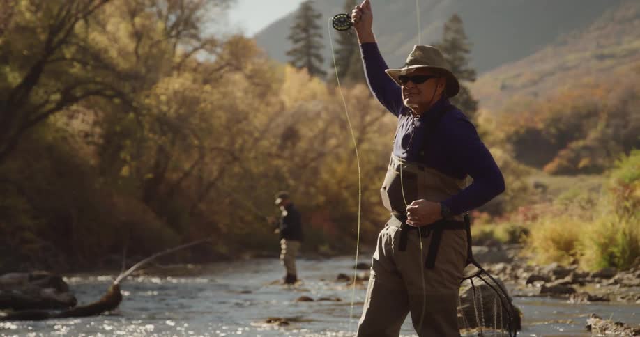 Two fishermen wearing waders fish in river winding past fall trees with misty mountain slope in the background