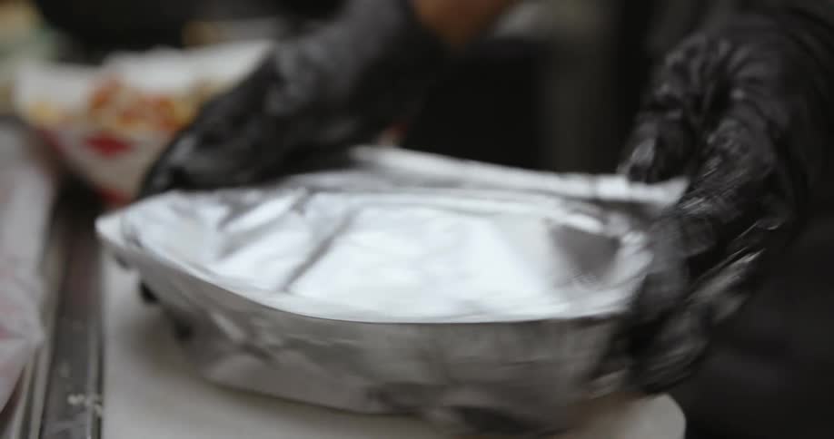 Woman wearing black latex gloves wraps take out food in aluminum foil