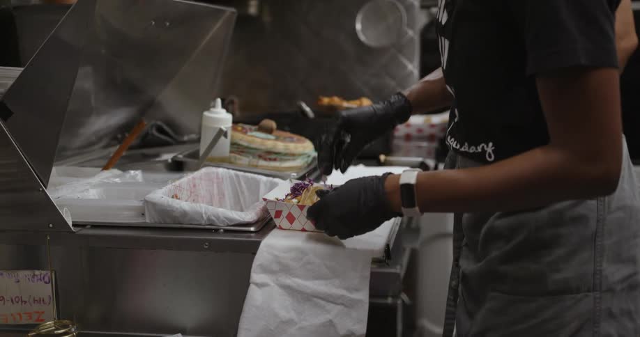 Woman with dreadlocks white headband black t shirt gloves and apron prepares fast food next to man frying chicken in the kitchen