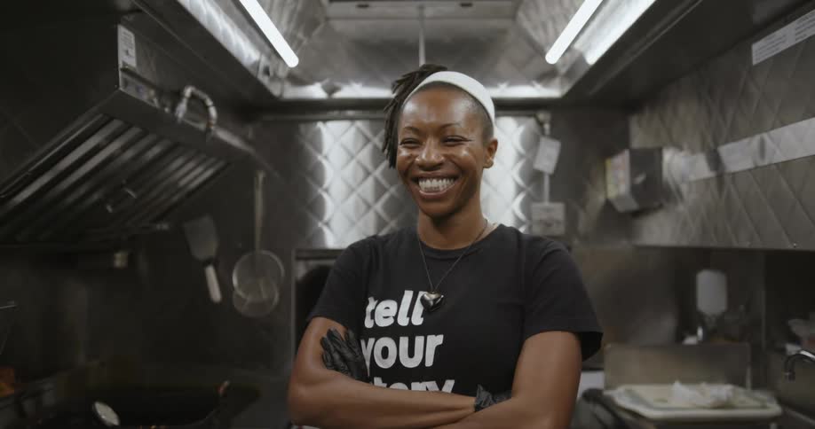 Female chef with dreadlocks white headband black t shirt necklace and gloves smiles broadly at camera in the kitchen