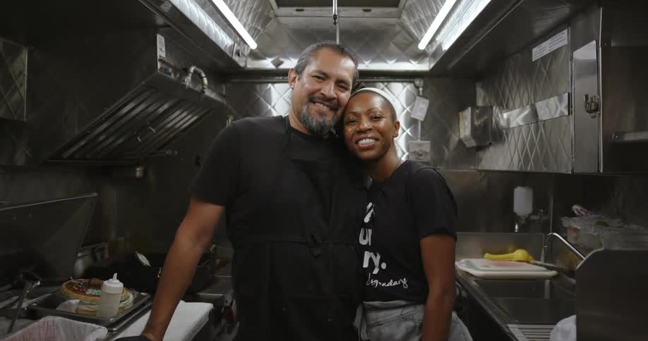 Man wearing black shirt and apron and woman in white headband black shirt and jeans smiles at camera in the kitchen