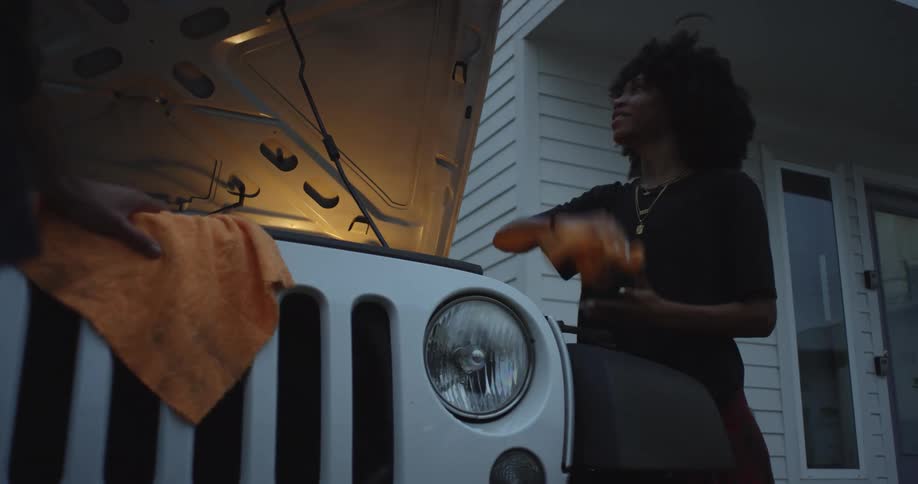 Woman with curly hair and black shirt cleans jeeps engine next to white building