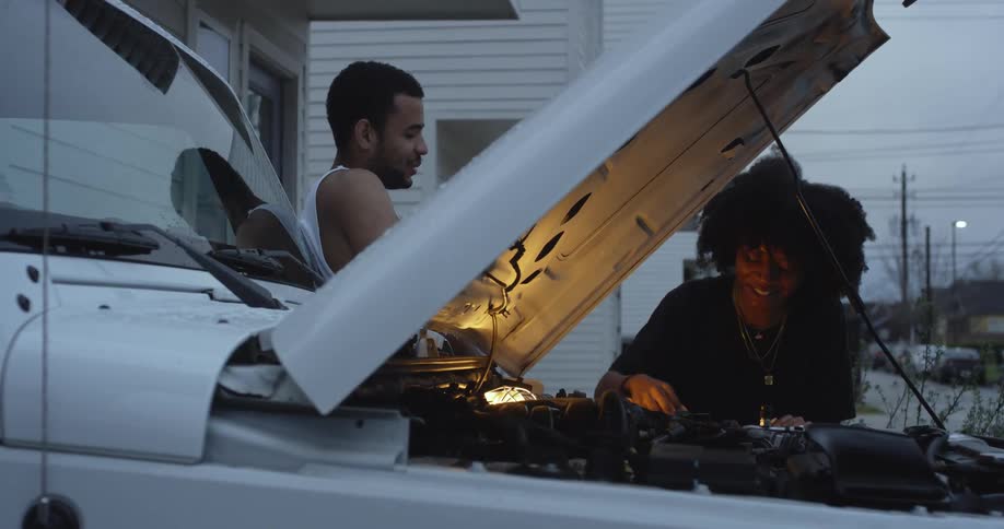 A man talks with a woman as she cleans the engine of a vehicle parked in the driveway of a house on a cloudy evening