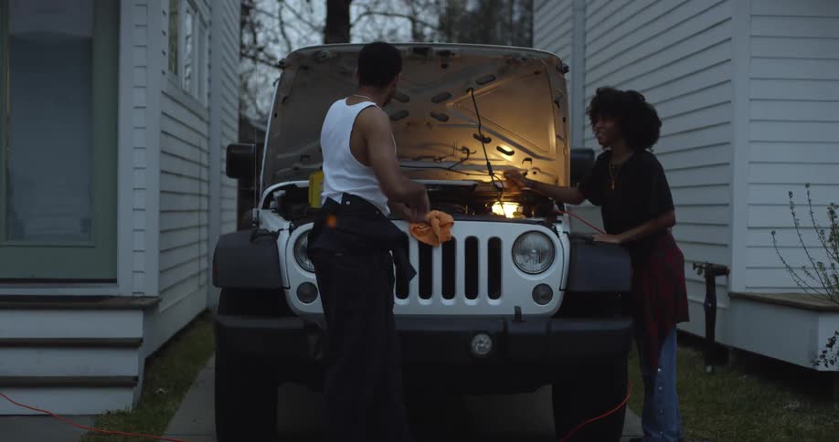 Man in white under shirt and black pants and woman with curly hair red shirt and jeans cleans white jeep engine with a piece of cloth between houses in the street