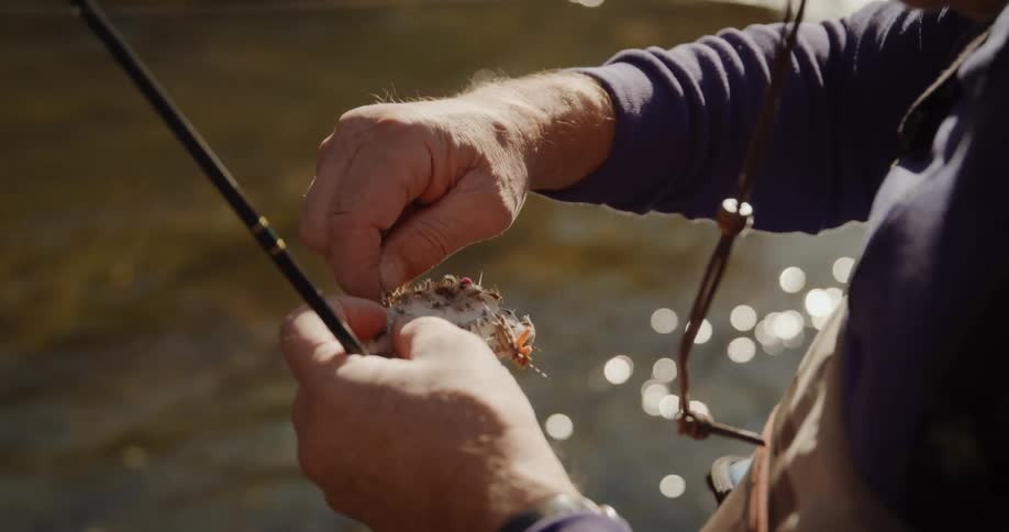 A man is standing near the river putting bait on his fishing line