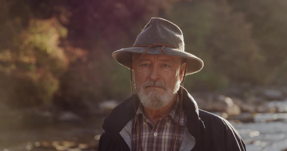 Elderly man with hat beard shirt and coat looks at camera near the stream in the autumn forest