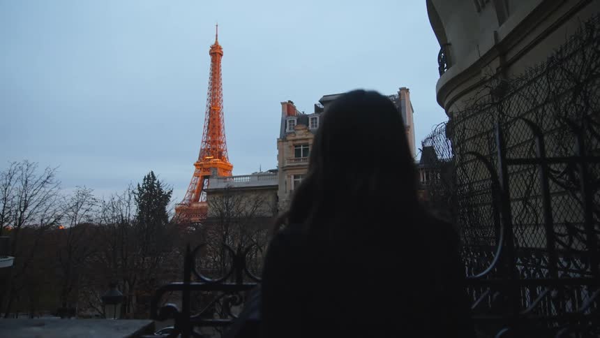 Woman with black coat and wavy hair walks by building and fences looking up at illuminated eiffel tower in paris